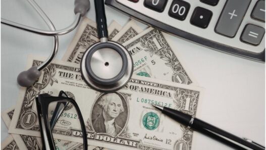 Calculator, stethoscope, dollar bills, pen, and eyeglasses on a desk