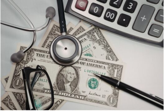 Calculator, stethoscope, dollar bills, pen, and eyeglasses on a desk