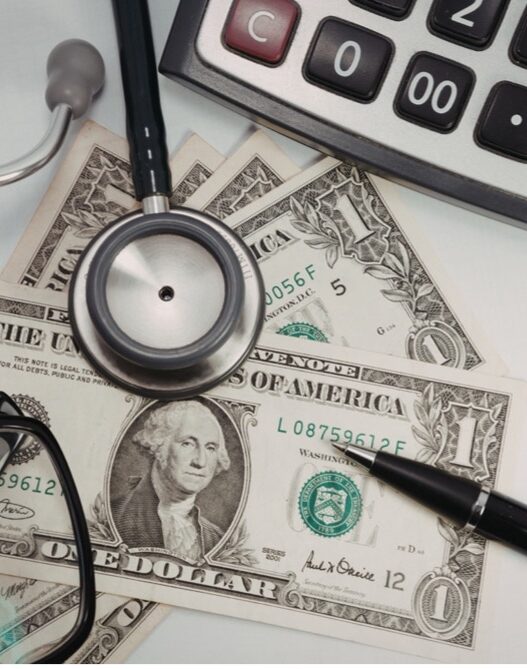 Calculator, stethoscope, dollar bills, pen, and eyeglasses on a desk