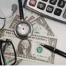 Calculator, stethoscope, dollar bills, pen, and eyeglasses on a desk